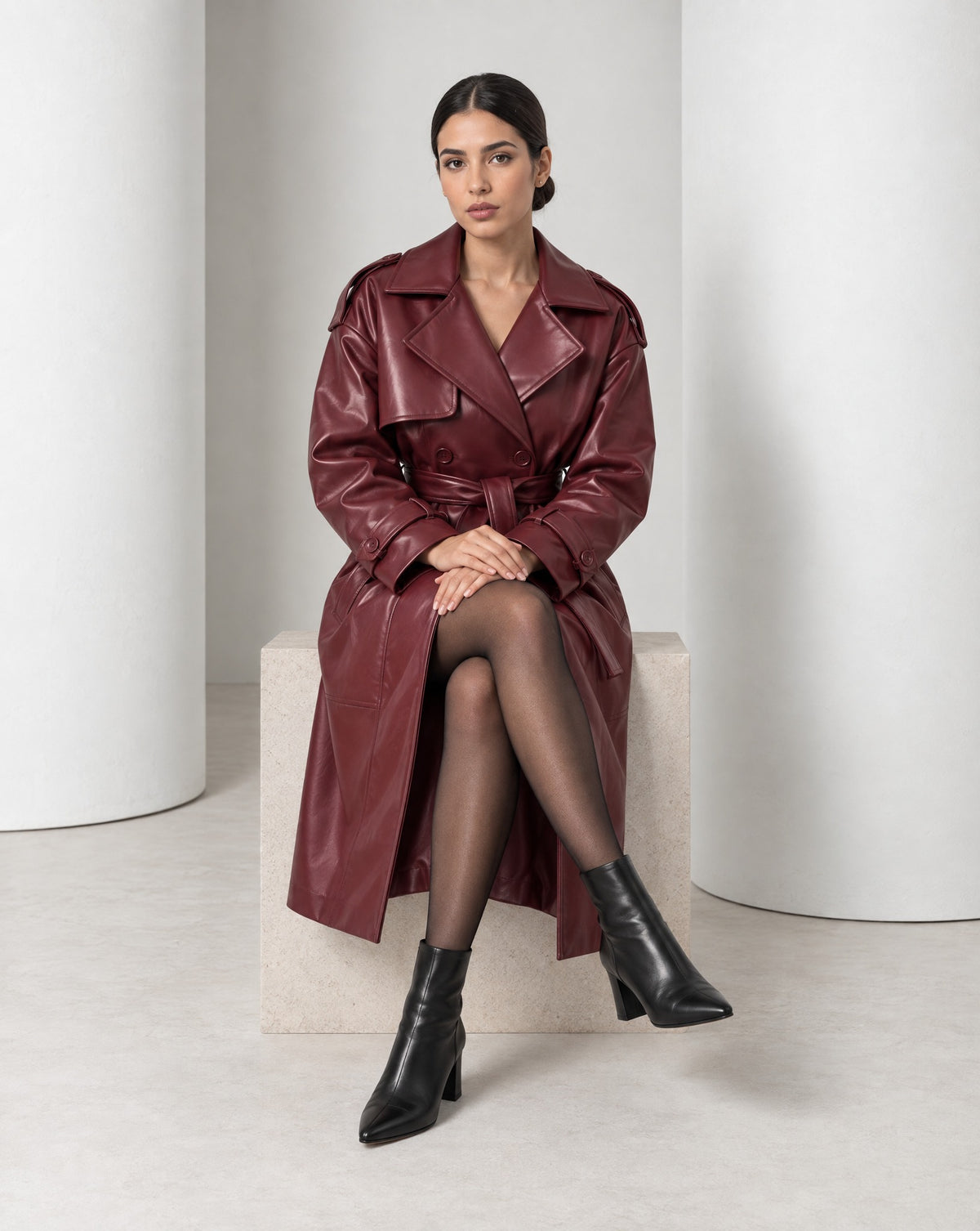 Model seated in the Alya burgundy trench, the coat belted and the skirt falling open over her legs
