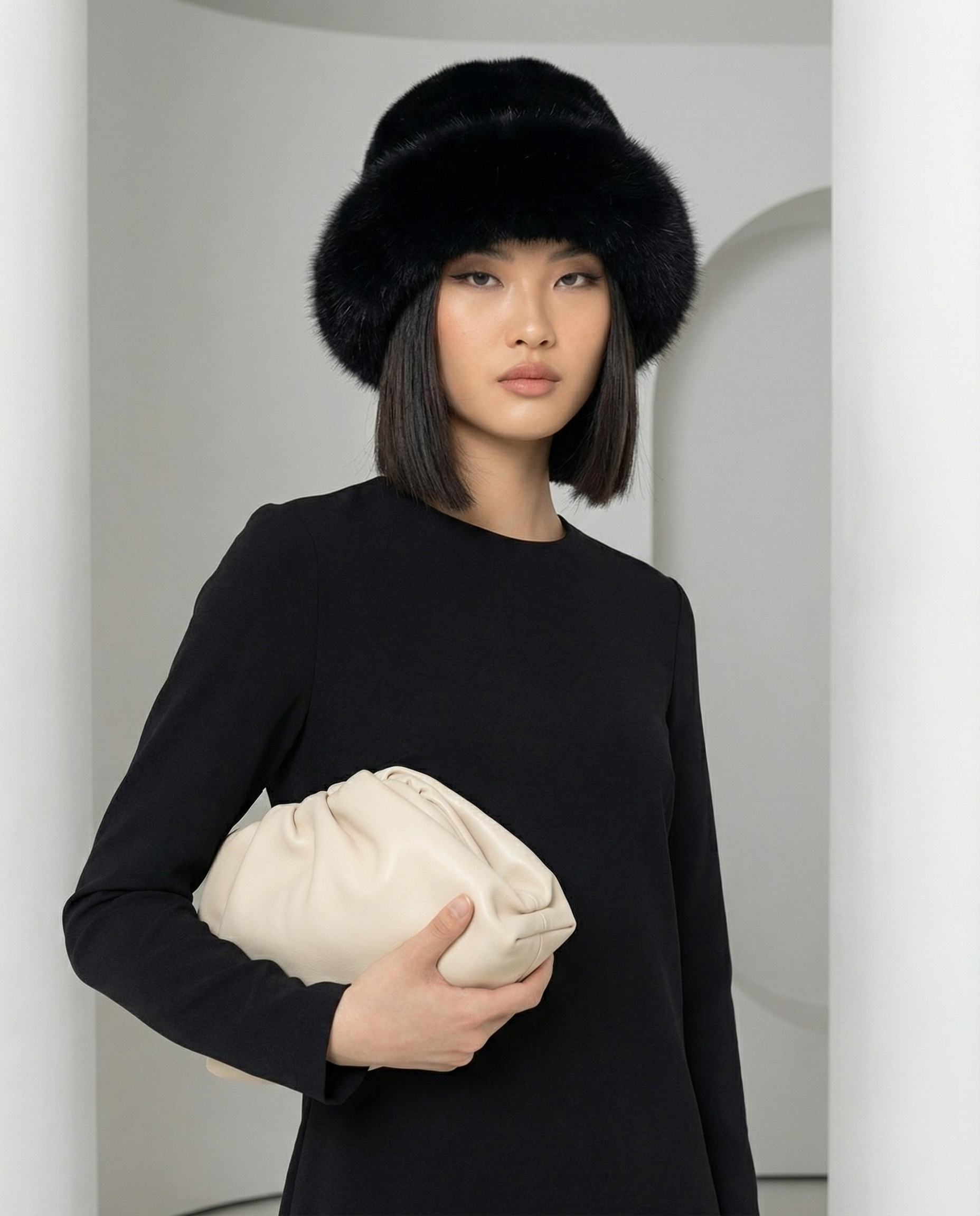 Black faux-fur bucket hat shot waist up over a long-sleeve black dress, cream clutch in hand
