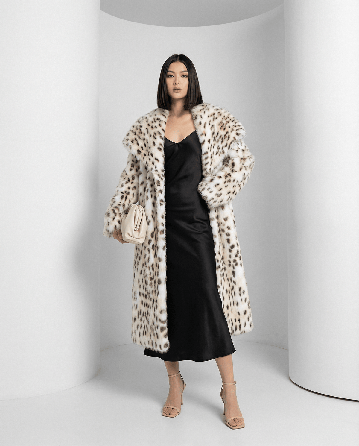 Snow-leopard faux-fur coat in a three-quarter turn over a black slip dress, cream clutch in hand