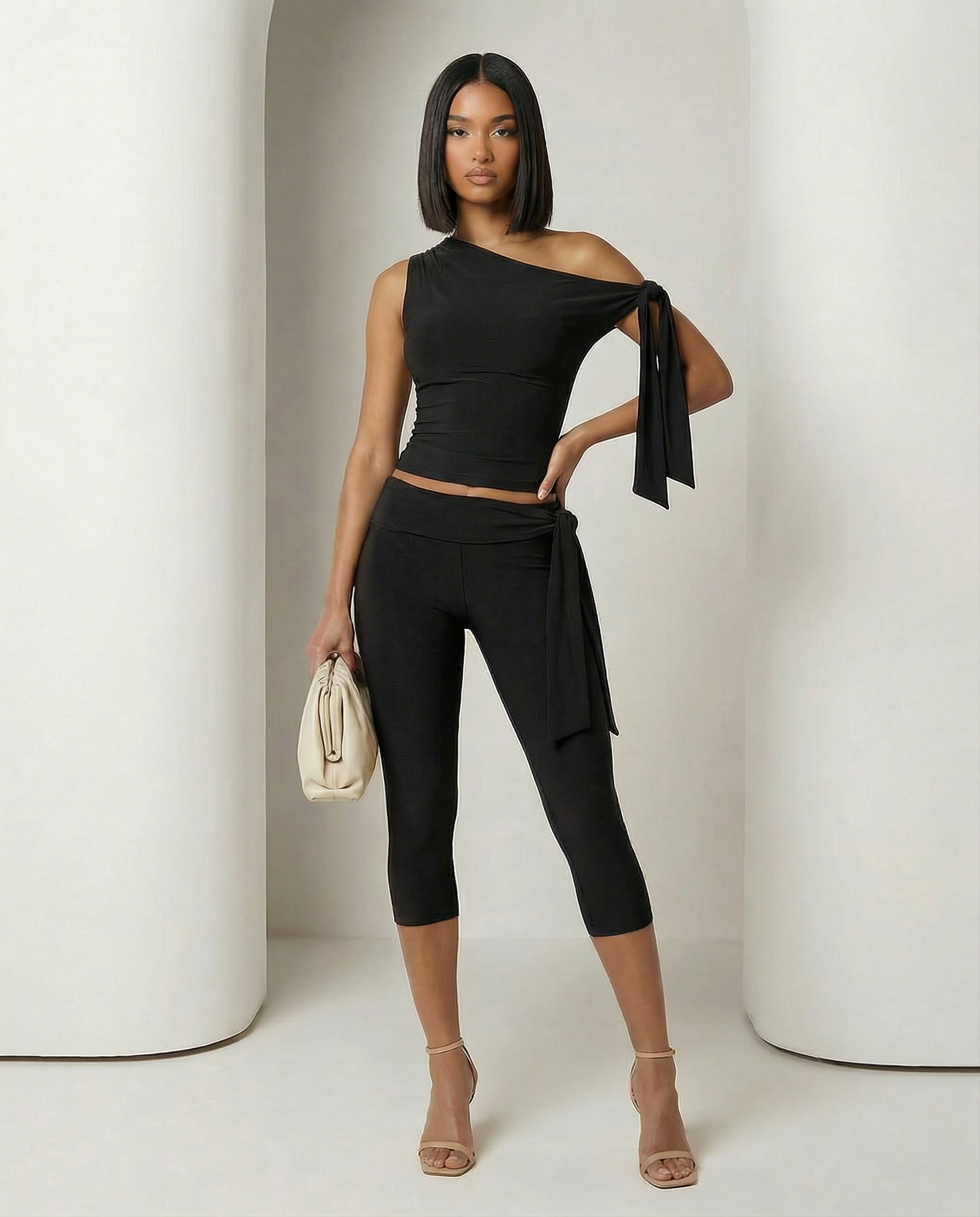 Black one-shoulder ruched top and capri leggings, hand on hip holding a cream pouch clutch