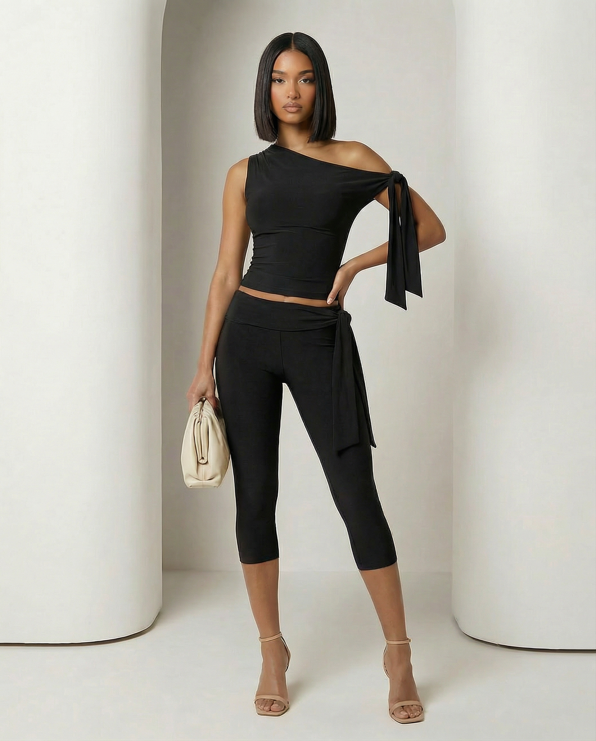 Black one-shoulder ruched top and capri leggings, hand on hip holding a cream pouch clutch