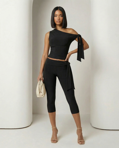 Black one-shoulder ruched top and capri leggings, hand on hip holding a cream pouch clutch