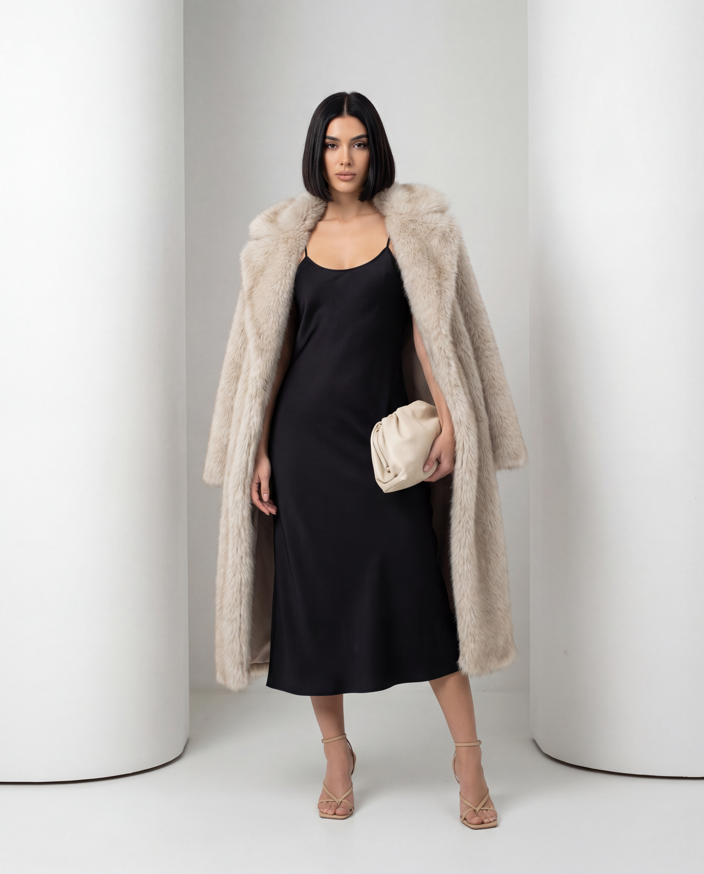 Mirella longline faux-fur coat in beige worn off the shoulders over a black slip dress, full length