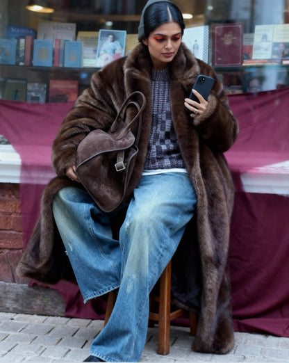 Bitter chocolate faux-fur coat over a checked knit and wide-leg jeans, seated outside a bookshop window