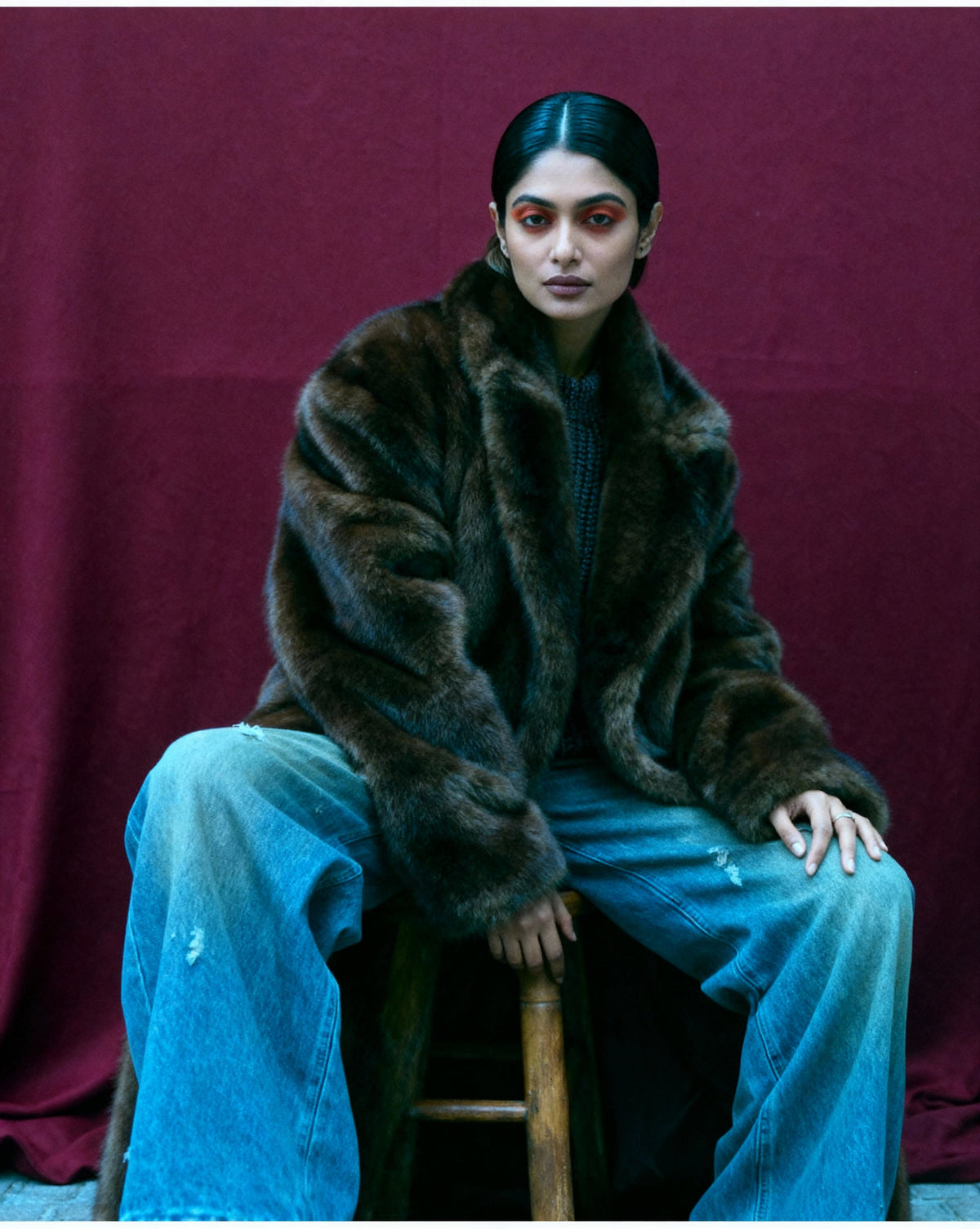 Bitter chocolate faux-fur coat with sleeves pushed back over a dark knit, seated against a burgundy drape