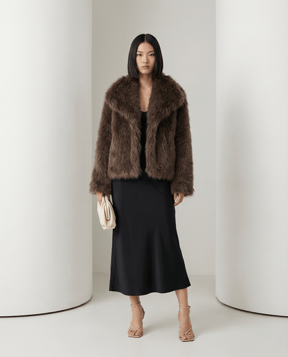 Chocolate brown cropped faux-fur jacket, three-quarter view with a cream clutch held at the hip