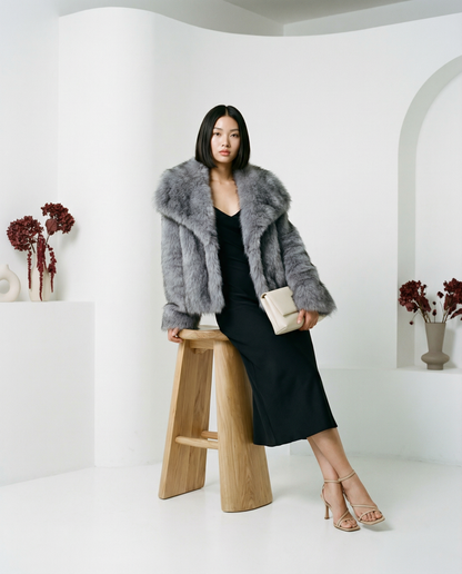 Gray Neriah plush faux-fur jacket worn leaning on a wooden stool beside a black midi dress