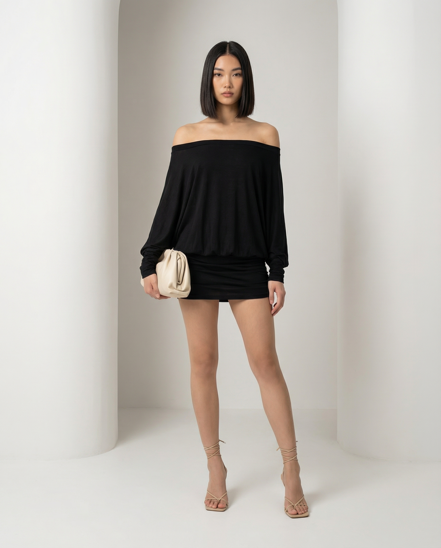 Black off-shoulder mini dress with dropped neckline and a cream clutch held at the hip