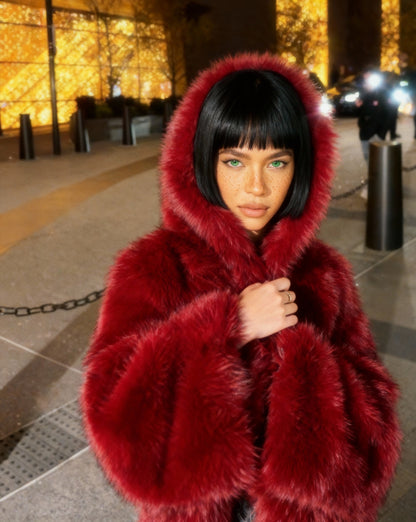 Scarlet red hooded faux-fur jacket with the hood up, waist-up night street shot with hands at the chest