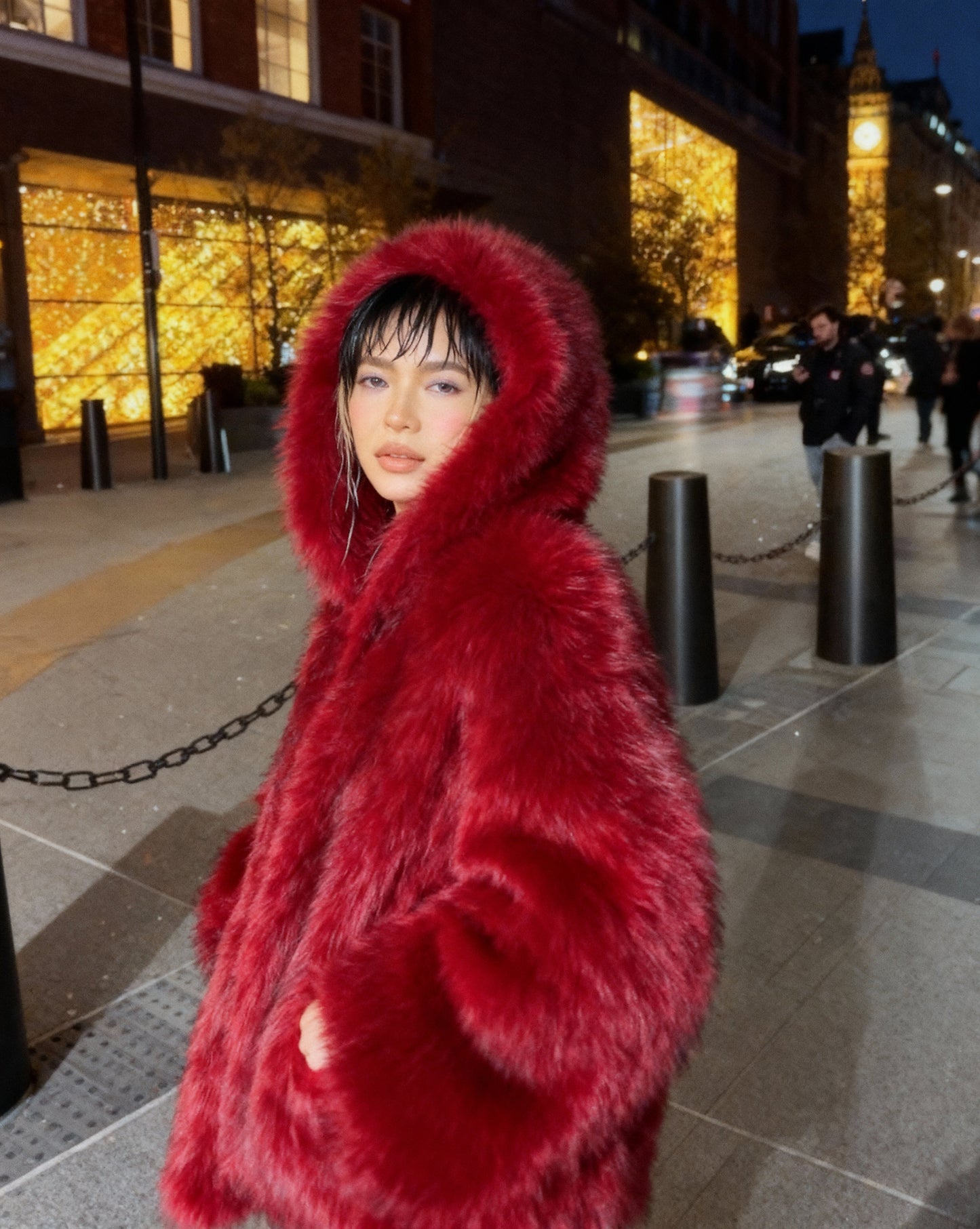 Scarlet red hooded faux-fur jacket with the hood up, three-quarter street view with hand in pocket