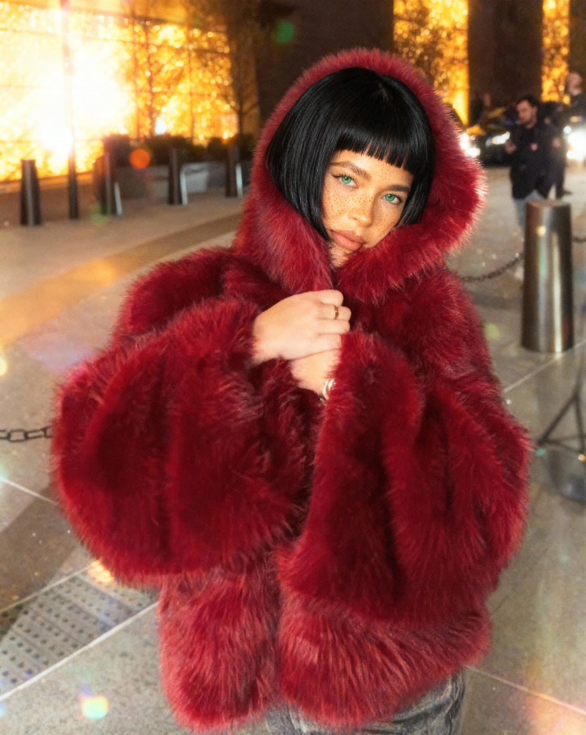 Scarlet red hooded faux-fur jacket clutched closed at the chest, hood up, waist-up night portrait