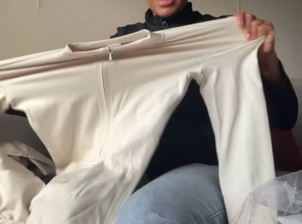 Unboxing shot of a cream long-sleeve zip-front jumpsuit held up by its shoulders on a sofa