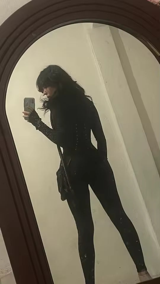 Side view of a black long-sleeve corset jumpsuit in an arched mirror, bag strap across the body