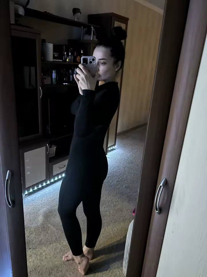 Back three-quarter view of a black long-sleeve jumpsuit in a dim living-room mirror, barefoot