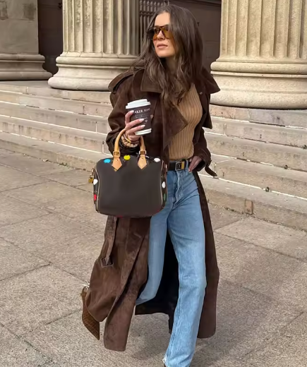 Brown suede trench worn open over a tan knit top and blue jeans, coffee in hand by stone columns