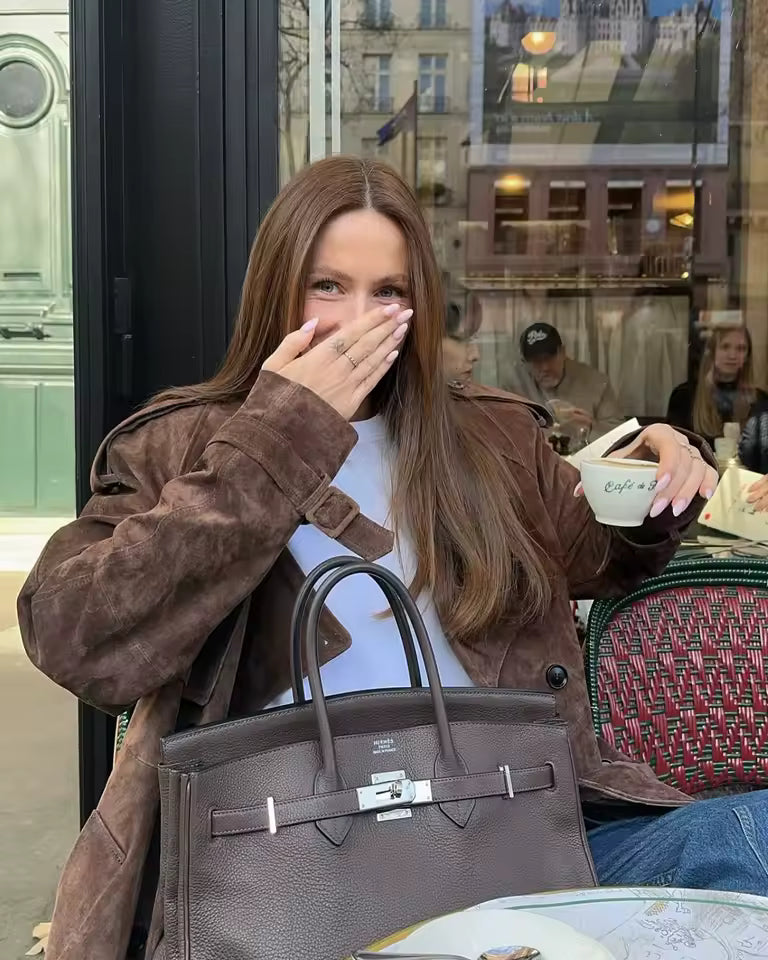 Brown suede jacket over a white tee, seated at a pavement cafe with a coffee cup
