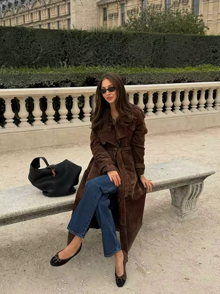 Long brown suede belted trench with jeans, seated on a stone bench in a formal garden