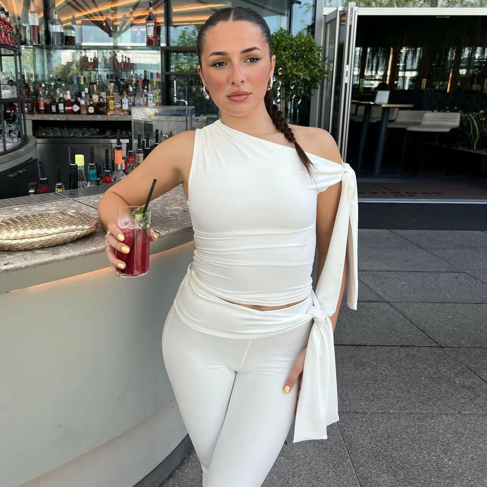 Leaning on a bar counter in a white one-shoulder top and slim white capris, holding a cocktail