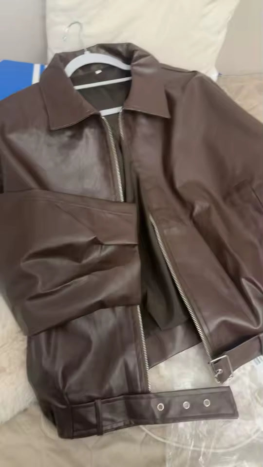 Brown leather jacket with a silver zip and belted hem, laid flat on a bed