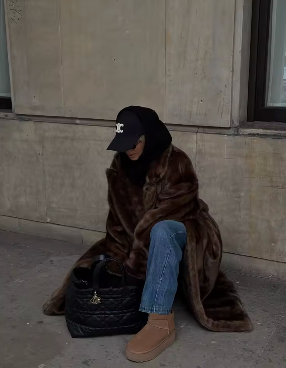 Long brown faux fur coat with jeans and platform boots, seated against a stone wall
