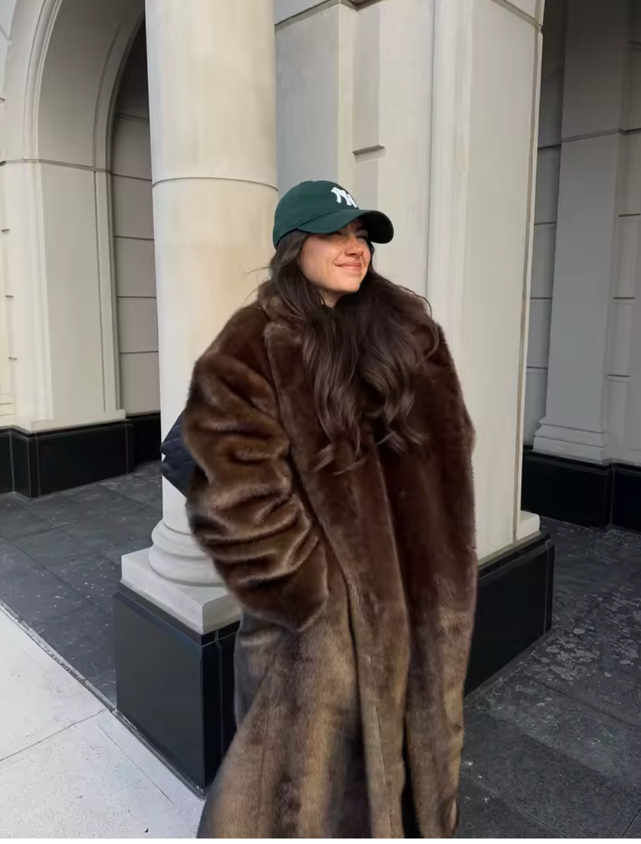 Brown faux fur coat worn open beside white stone columns, with a green baseball cap