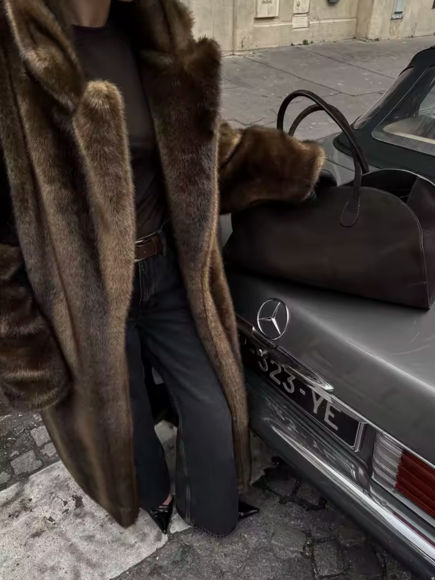 Full-length brown faux fur coat with jeans, beside a vintage car and a brown holdall