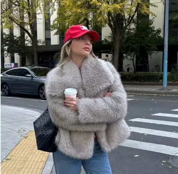 Grey faux fur cropped jacket worn open over jeans with a red cap and coffee cup at a city crossing