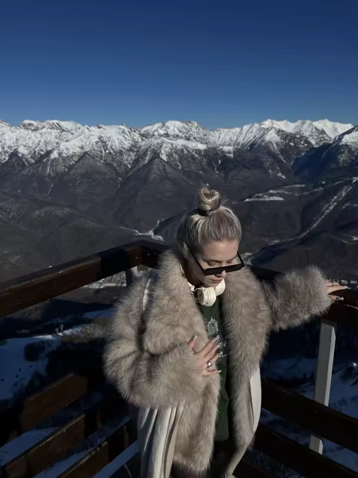 Taupe faux-fur coat worn open over a green tee at a snowy mountain viewpoint, sunglasses on