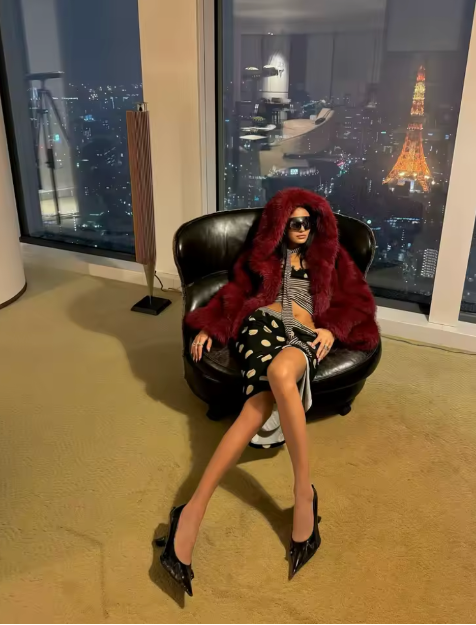 Deep burgundy hooded faux fur coat worn seated in a leather chair by a night skyline window