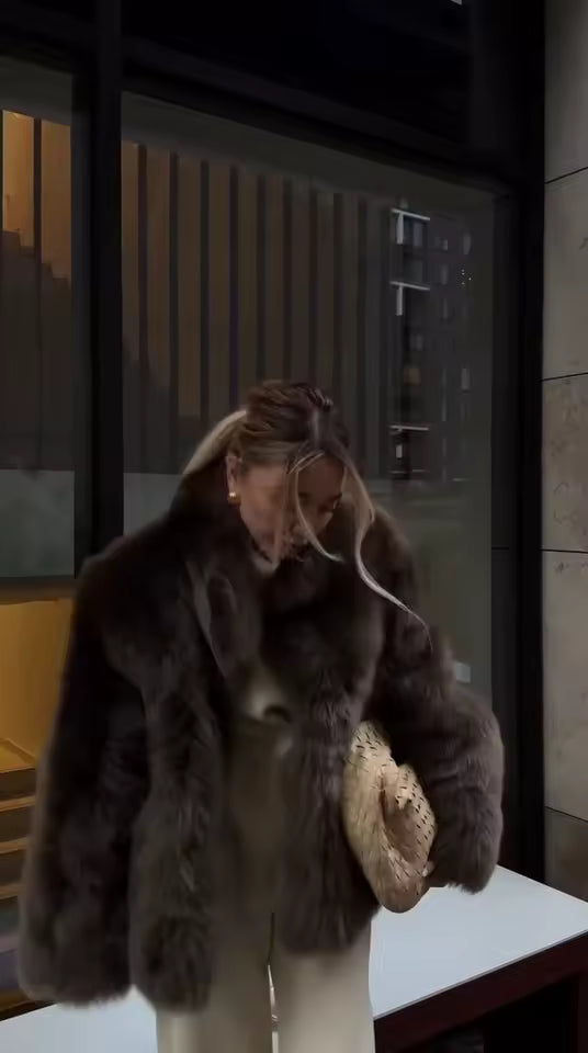 Deep brown faux-fur jacket over cream trousers with a woven clutch, shot outside at dusk