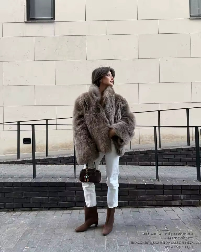 A woman in a grey-brown faux-fur jacket with white jeans and brown boots on a stone-walled street