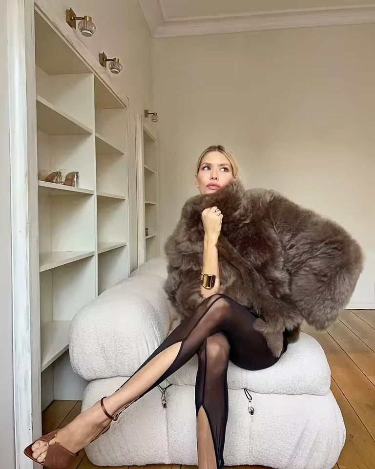 Taupe faux-fur jacket held around the shoulders over a black dress, seated on a boucle chair