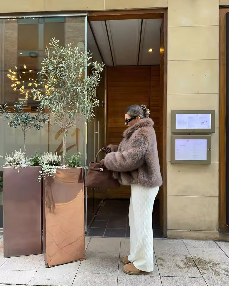 Mauve-taupe faux-fur jacket with cream knit trousers and a brown bag beside a doorway