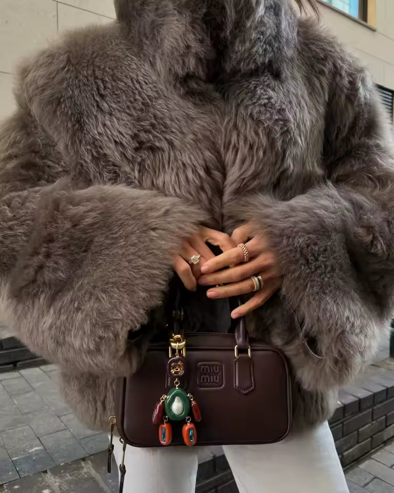 Waist-up crop of a shaggy grey faux-fur coat, ringed hands resting on a burgundy shoulder bag