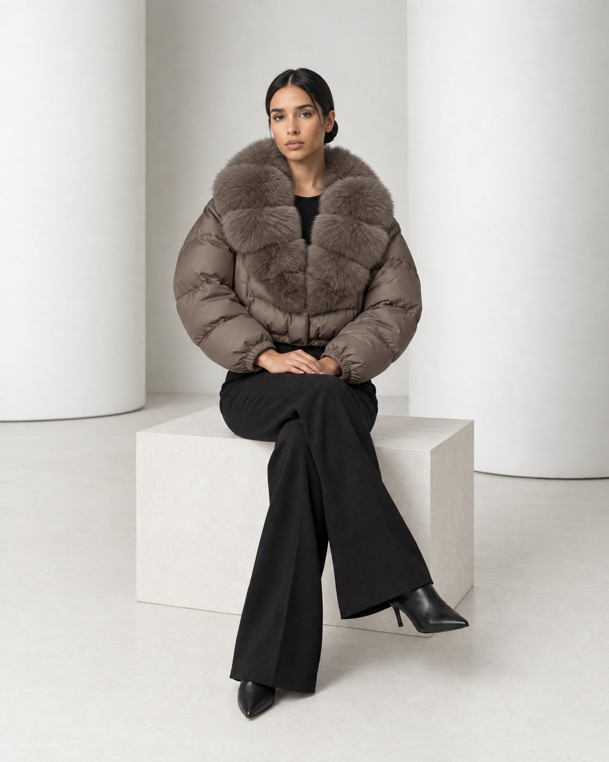 Model seated in the Saga taupe faux-fur collar puffer, the jacket closed and the cropped hem at her waist