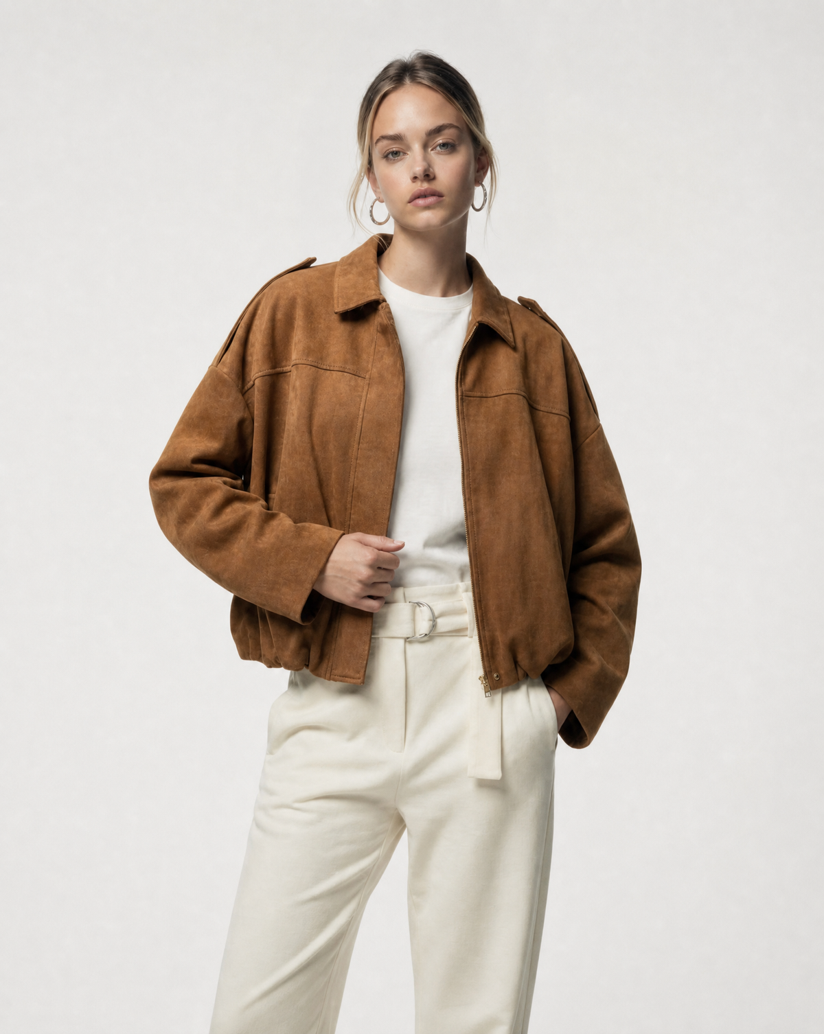 Tan suede bomber worn open with hands in the pockets over cream belted trousers, front view