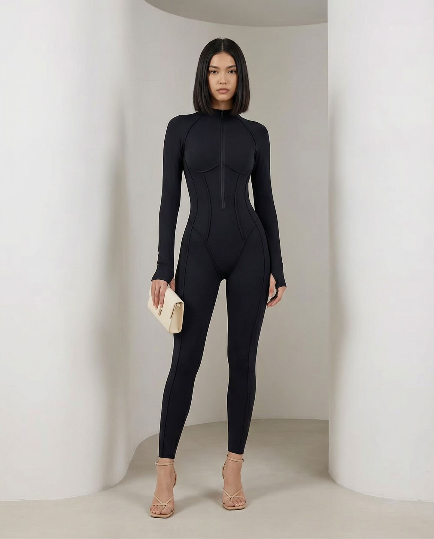 Selene Sculpt Jumpsuit in black with zip neck and contour seams, front view on a curved set