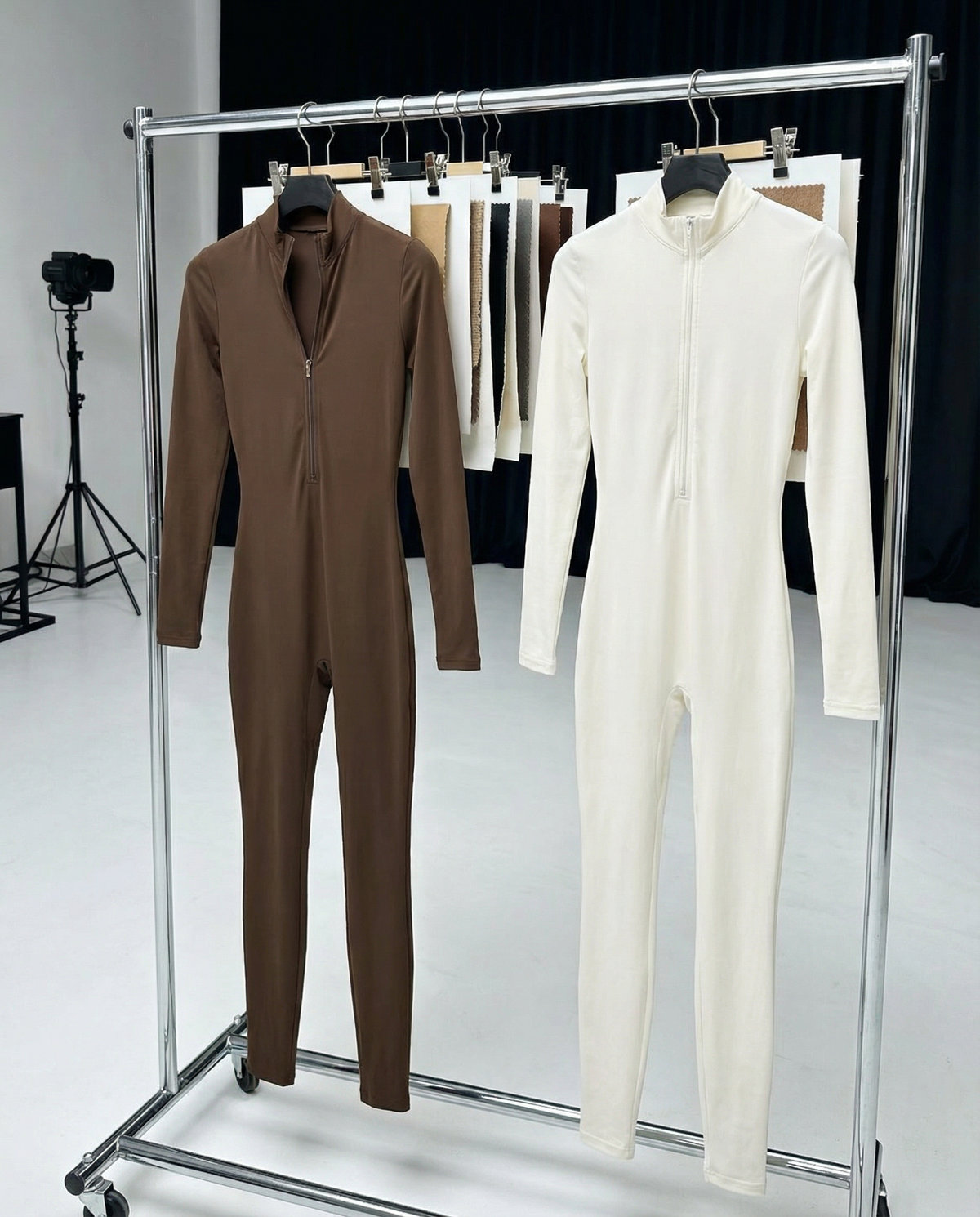 Brown and cream zip-front unitards hanging on a chrome rail in a studio, swatches behind