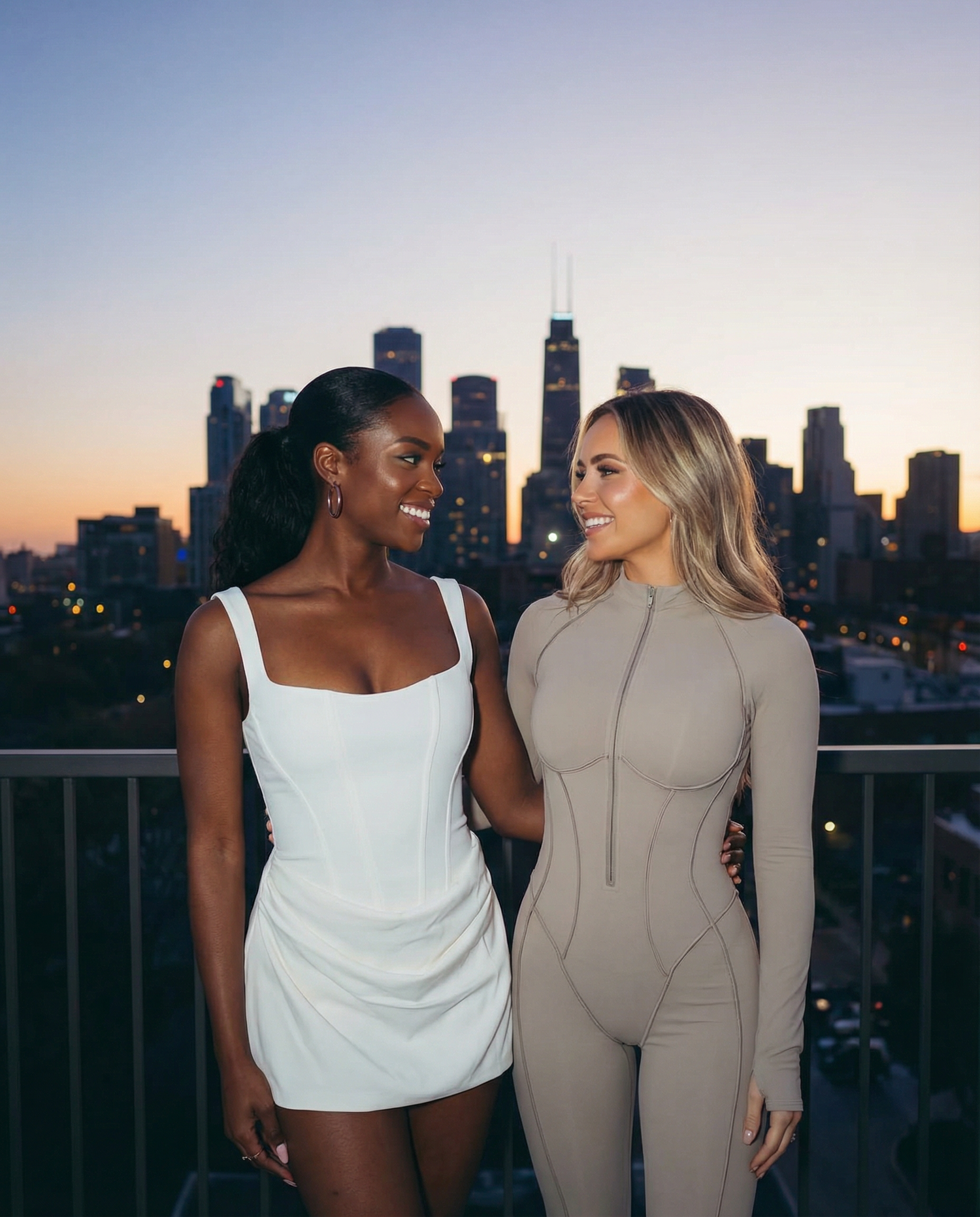 Two women on a rooftop at dusk, one in a white mini dress, one in a taupe zip unitard