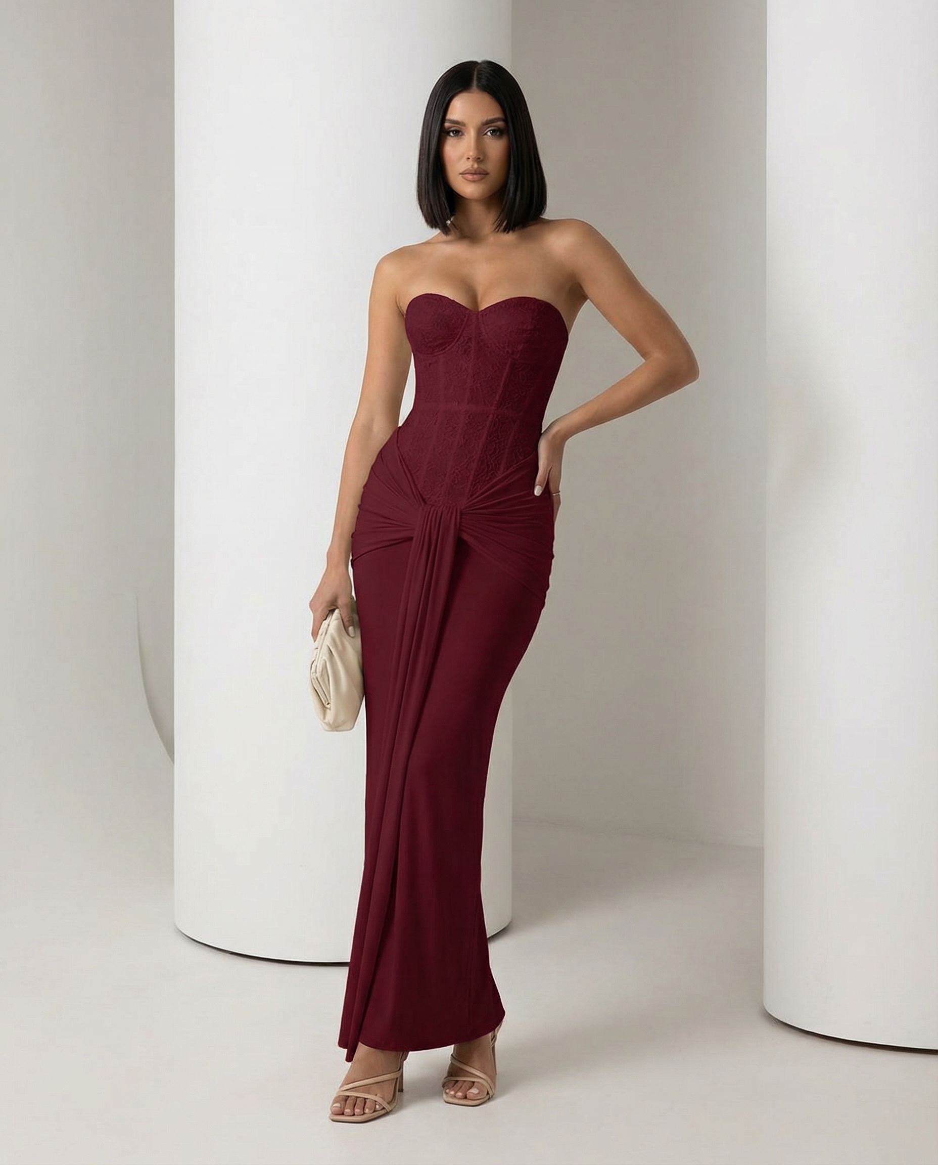 Burgundy strapless corset maxi dress with a knotted drape at the hip, hand on hip, full length
