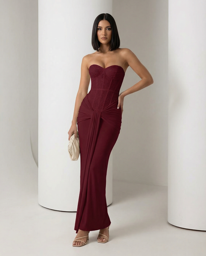 Burgundy strapless corset maxi dress with a knotted drape at the hip, hand on hip, full length