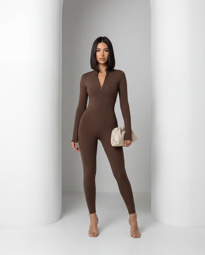 Skadi jumpsuit in bitter chocolate, standing between white columns with the zip pulled partway down