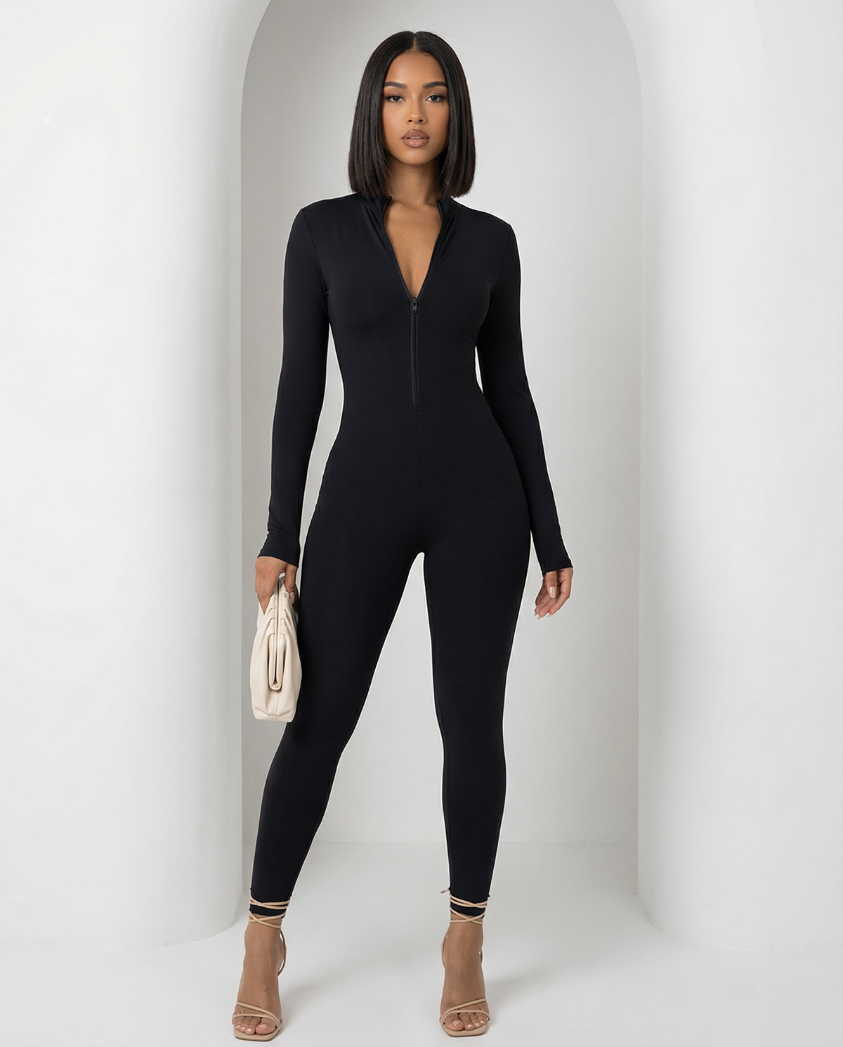 Black Skadi jumpsuit with half-zip mock neck, full length front view with a cream clutch